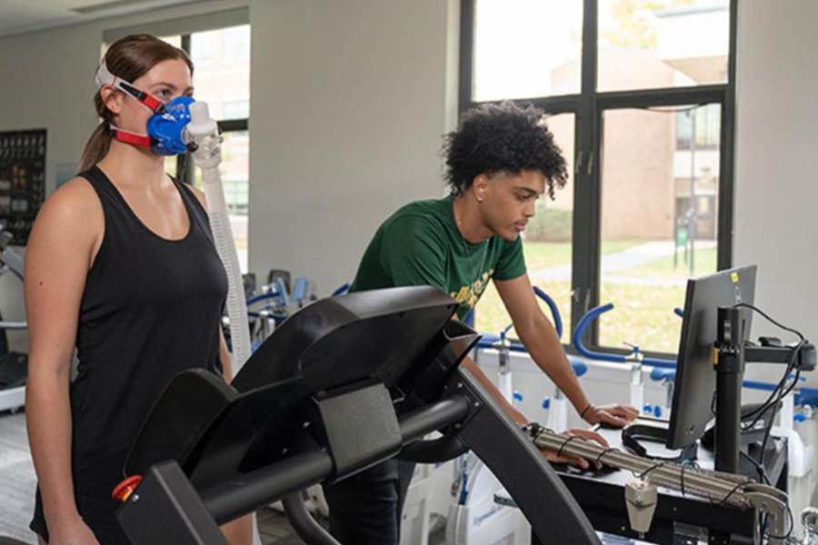 A student in a sports science lab monitors data on a computer while an athlete wearing a VO2 max respiratory mask runs on a treadmill.